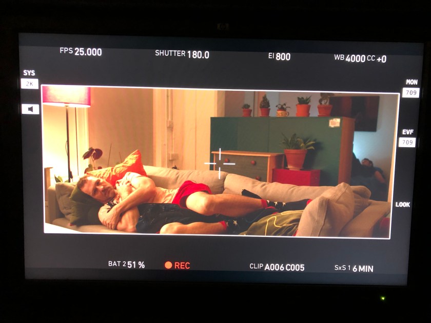 A man lies on the sofa hugging himself while in a separate alcove another person sleeps.