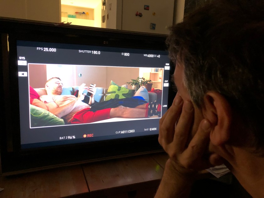 A man watches the monitor on which another man lies on the sofa holding his phone.