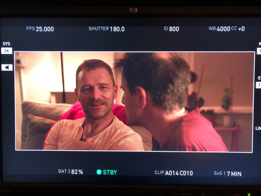 Two men in conversation, the one facing us, bearded, is smiling.