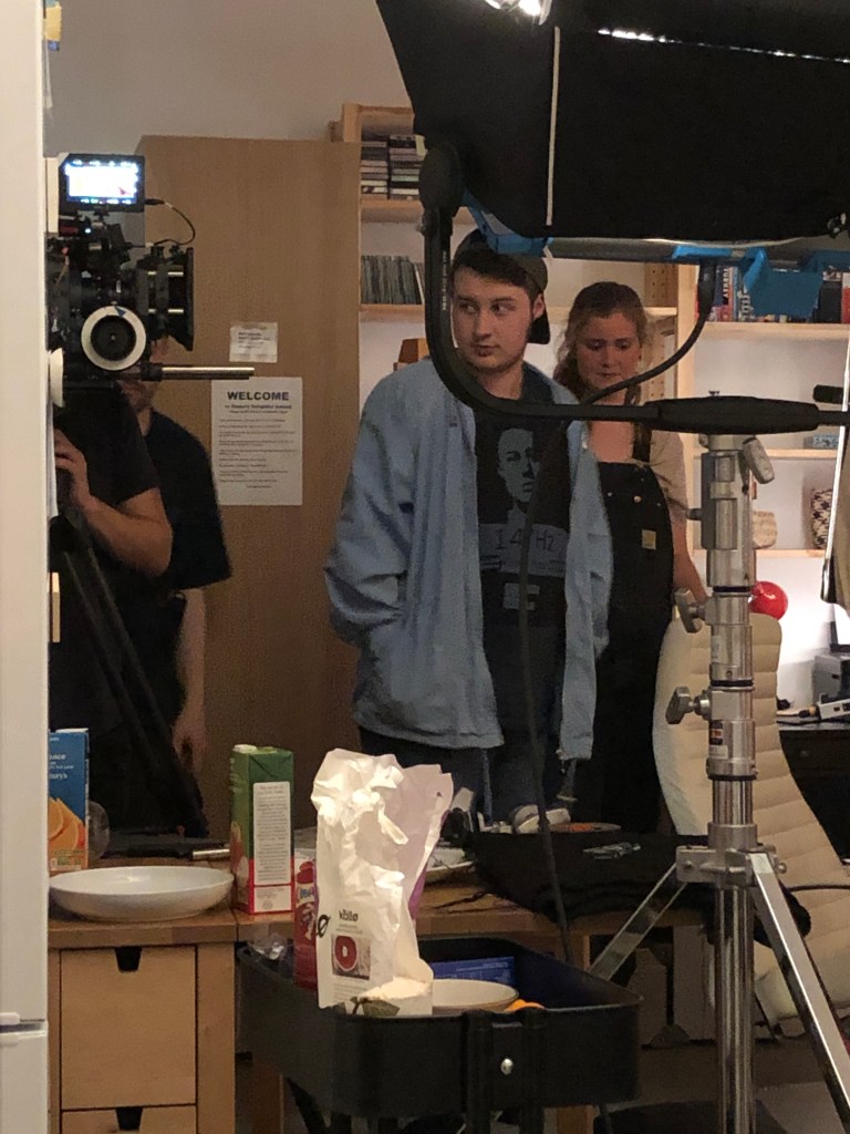An actor and electrical department member stand behind a light and camera.