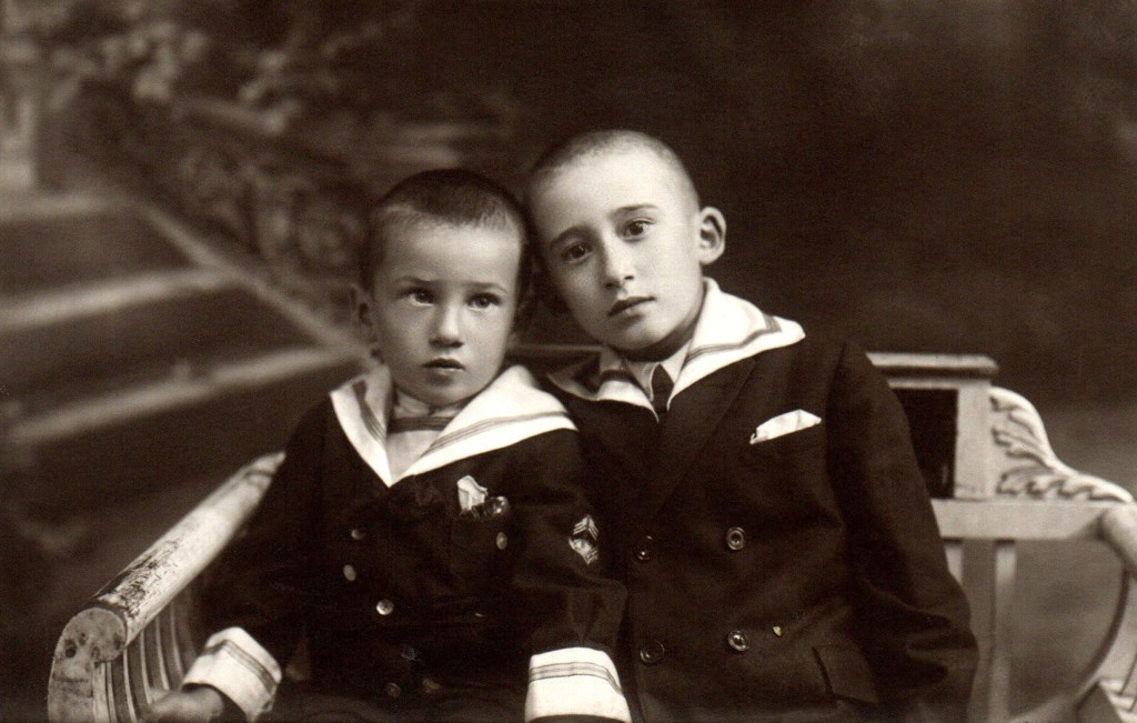 A sepia coloured portrait shows two young boyswearing sailor suits, seen from the waist up, their heads leaning against each other