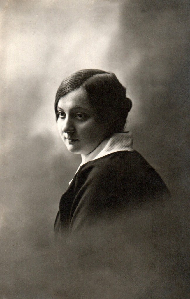 A sepia coloured portrait of a light skinned young woman seen from the shoulders up, her body faces backwards with her head turned towards us in three quarter profile.