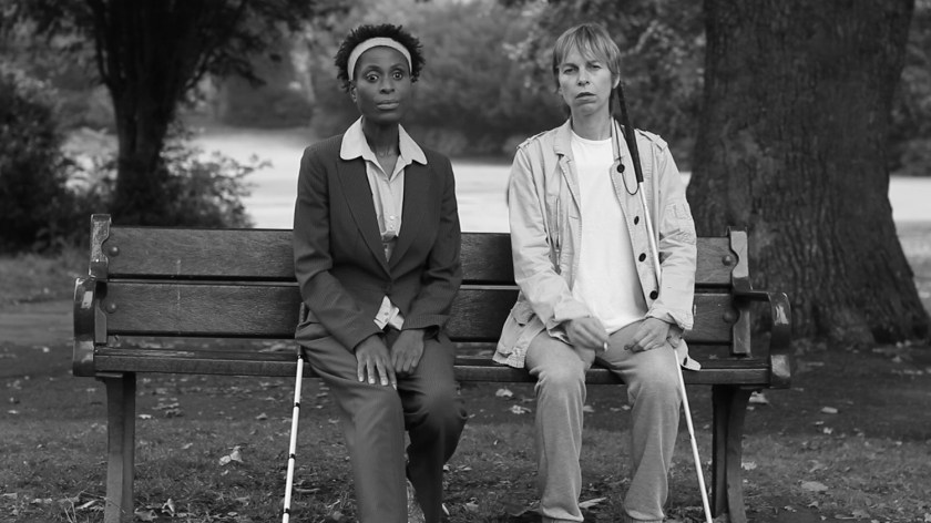 Black-and-white still from Awake: Doreen and Anna sit side by side on a park bench, facing forward, with their white canes resting beside them.