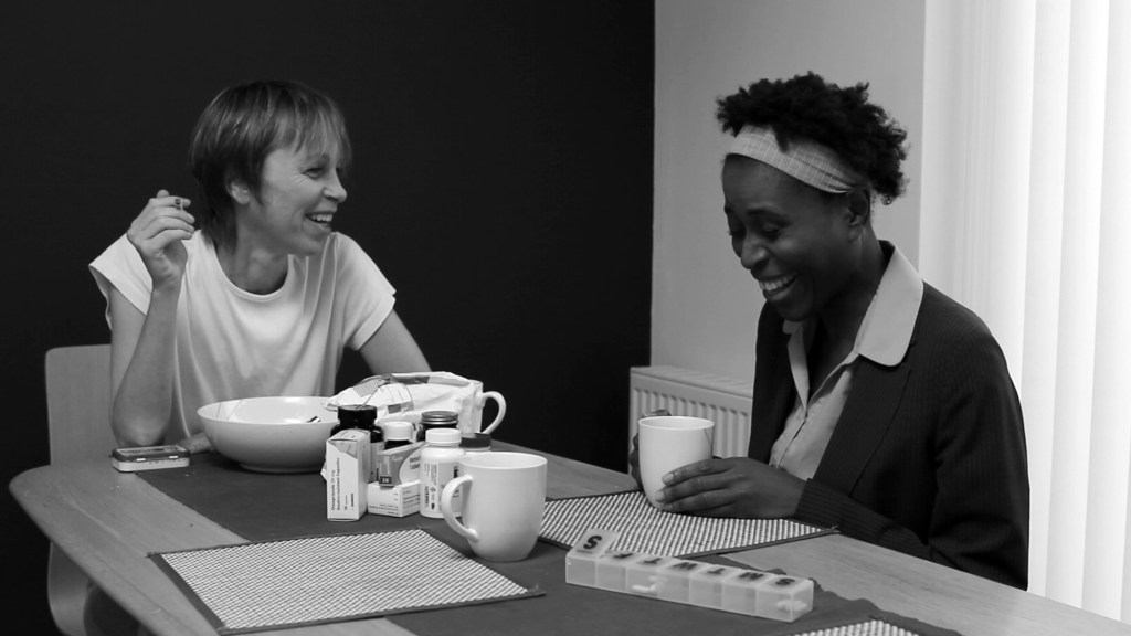Black-and-white still from Awake: Anna and Doreen sit laughing together at a kitchen table with mugs, food and medication between them.