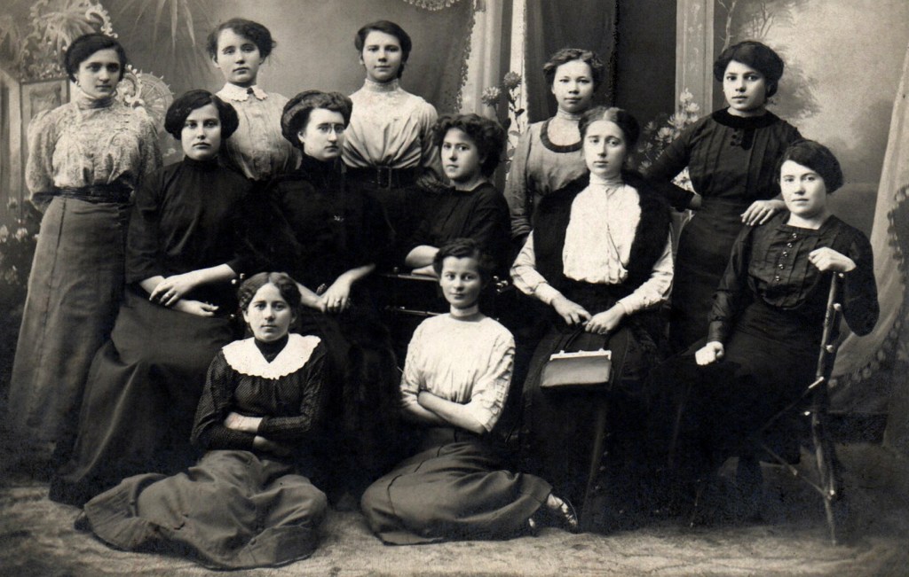 Sepia group portrait of eleven young women posed in three rows, wearing long dresses with high collars.
