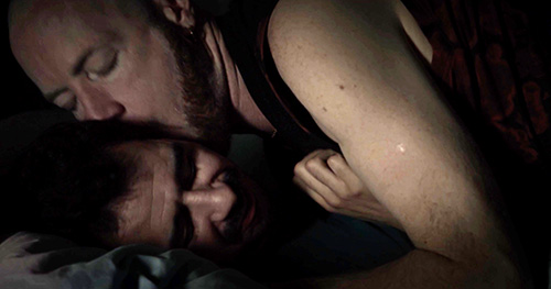 Film still from sift.simmer.shred: Arthur and Xavier lie closely together on a bed in a dimly lit room, one embracing the other from behind.