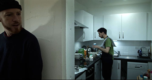 Film still from sift.simmer.shred: Arthur stands in the foreground of a small flat while Xavier prepares something at a cluttered kitchen counter behind him.