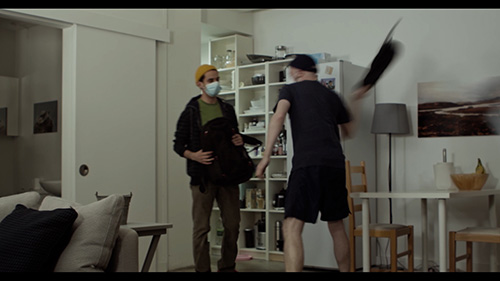 Film still from sift.simmer.shred: Xavier wearing a face mask defends himself with his rucksack as Arthur, seen from behind, moves to thrash him with a coat.